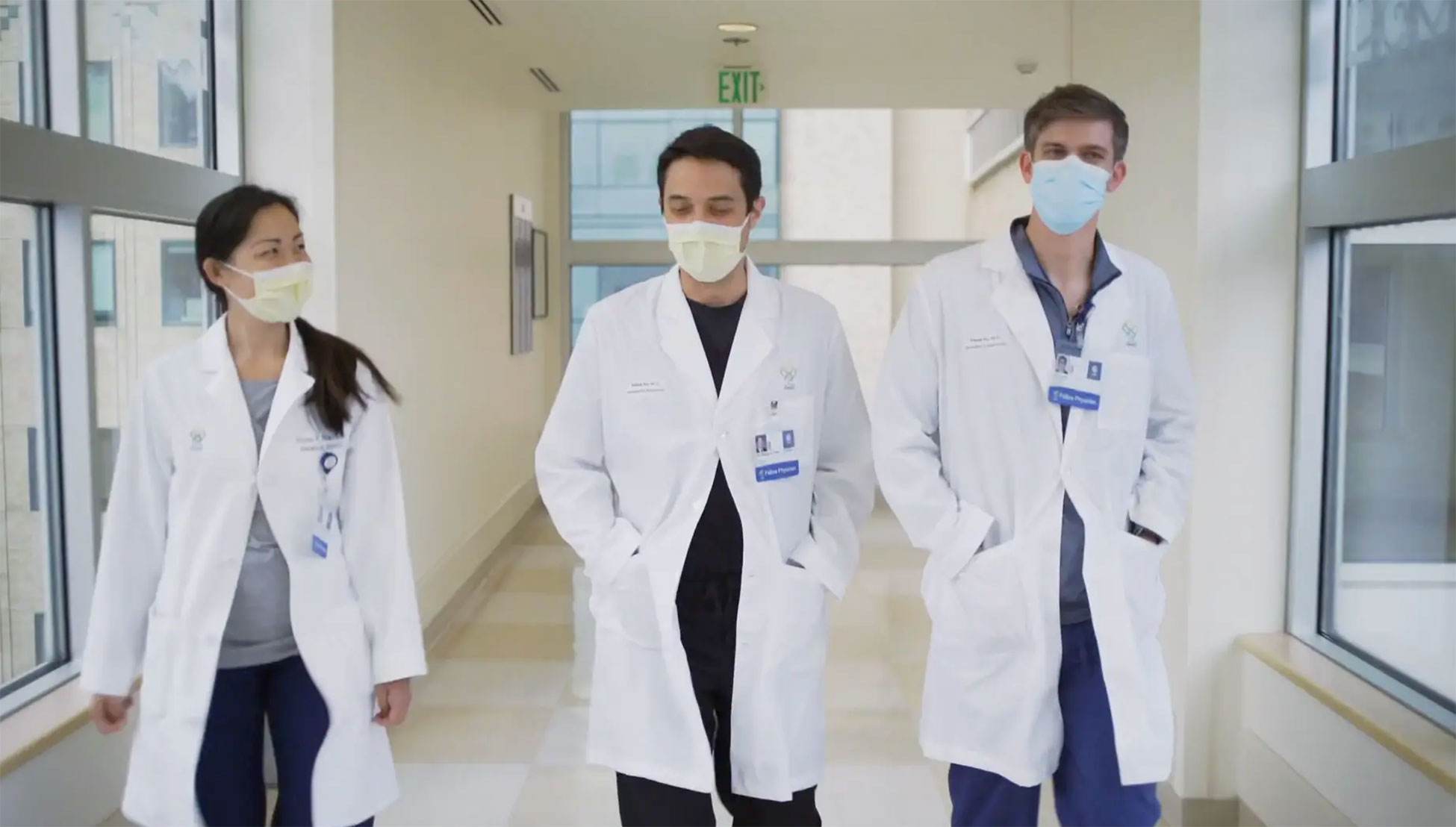 Three medical students in white coats wearing face masks walk down a hallway.