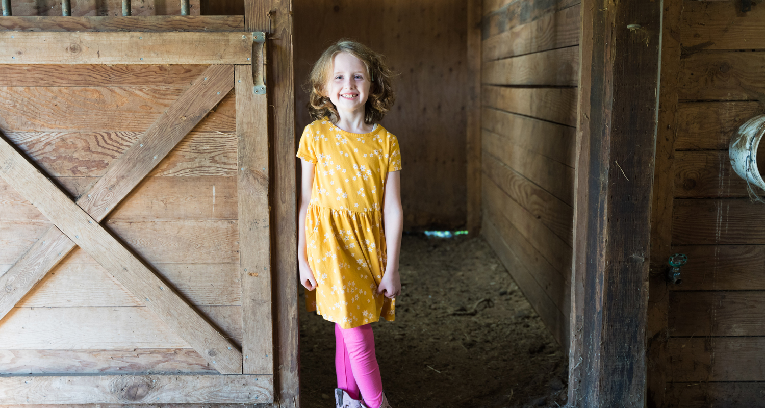Little girl grins, standing at a barn door