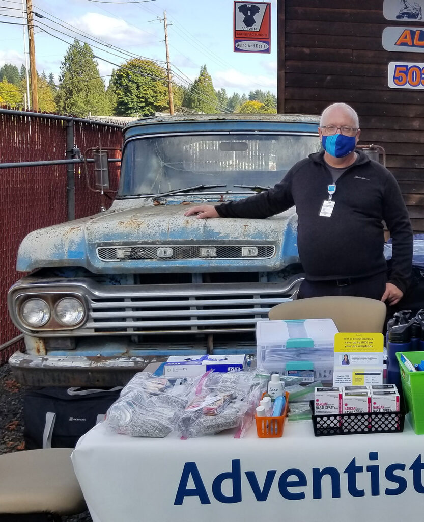 Older white man stands next to a Ford truck, in front of a table of community health supplies