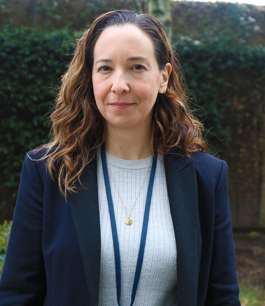 A woman wears a gray sweater and navy blazer, with a slight smile on her face