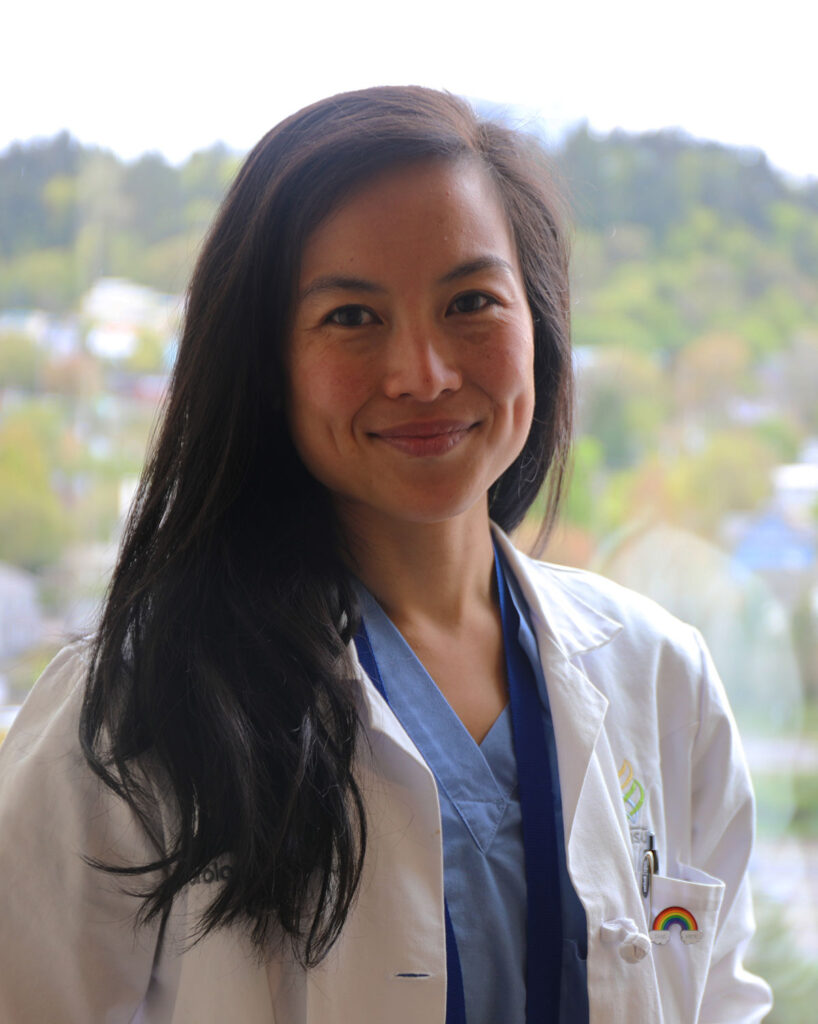 Surgeon with long black hair, blue scrubs and white lab coat smiles by window