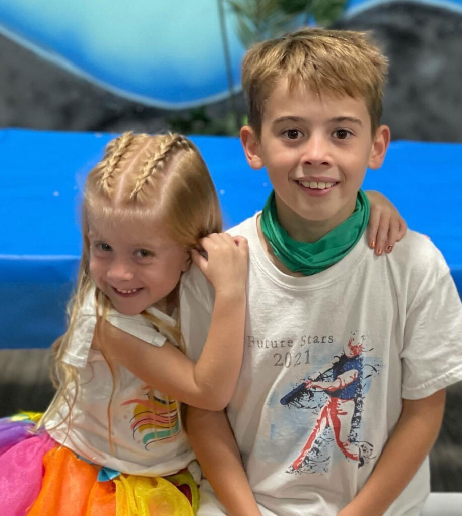 A little girl in a rainbow tutu and her brother sit next to one another, smiling in