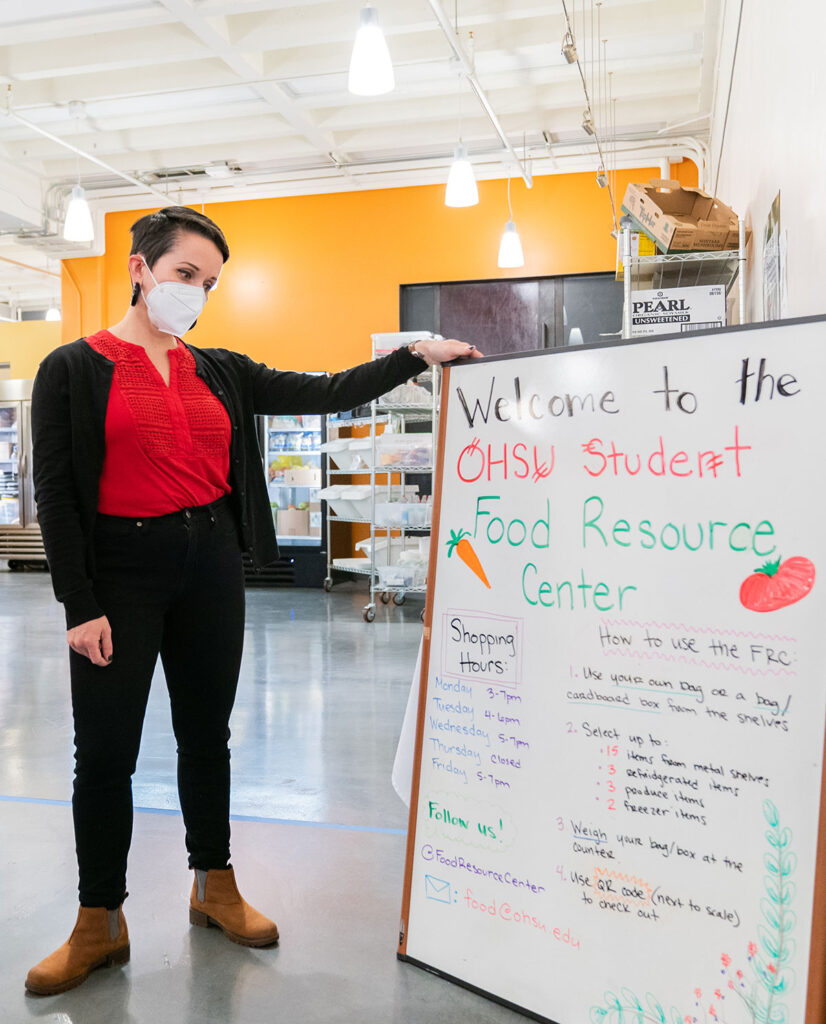 A woman with short dark hair in a red shirt and black trousers holds a whiteboard sign with instructions on how to use the Food Resource Center