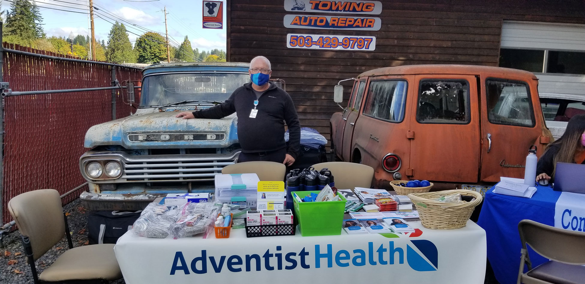 A nurse stands at a table at a community event with an auto shop in the background