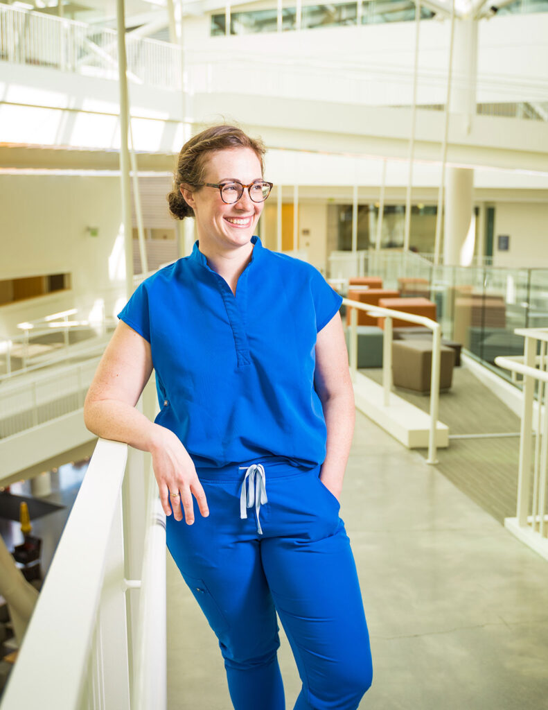 Woman with brown hair, glasses and blue scrubs leans against the railing inside the Robertson Life Sciences Building
