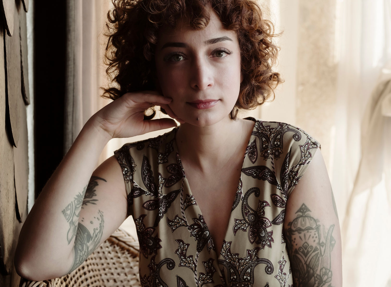 Young white woman with curly auburn hair and tattoos on her arms looks defiantly at the camera