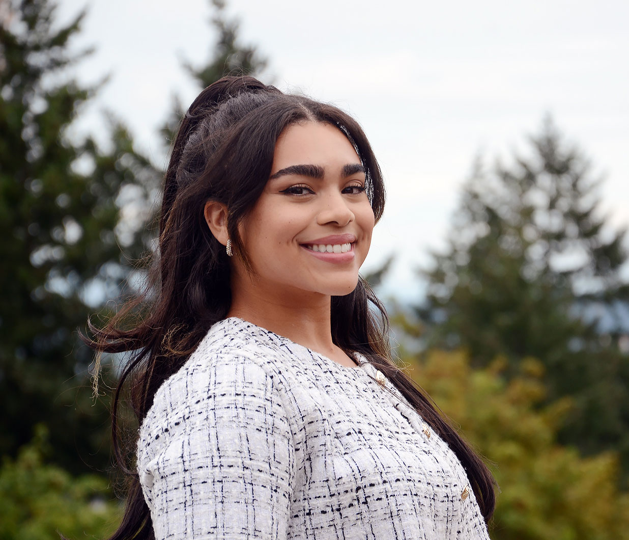 A dark haired young woman smiles at the camera. She is outside with blurred trees in the background