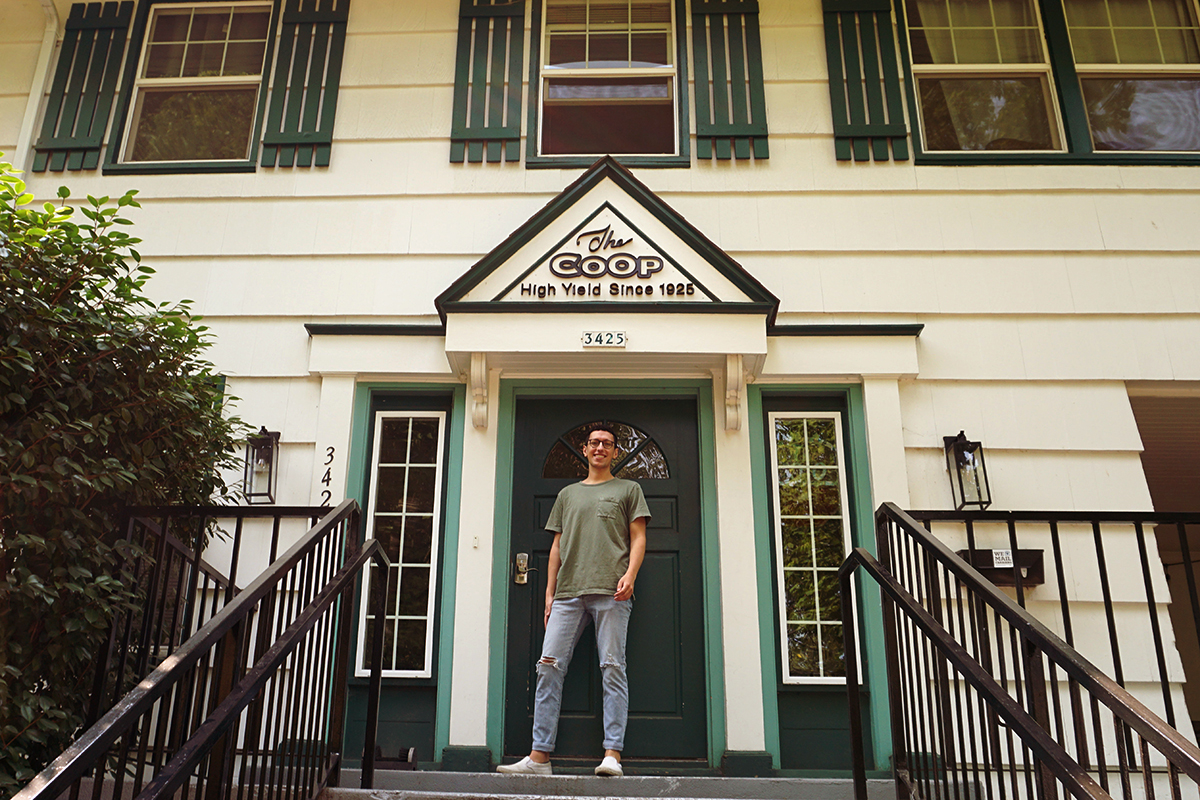 A young man in jeans and a t-shirt stands on the steps of a white two story house with green shutters and a sign above the door that says "The Co-op High Yield Since 1925"