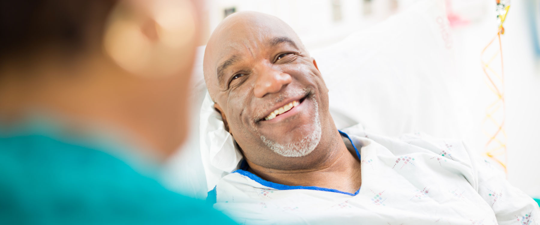 A man in a hospital bed, wearing a gown, smiles at a person who's blurred in the foreground