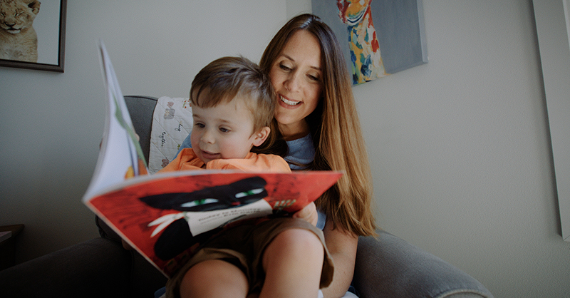 A mother reads to her toddler, sitting on her lap.