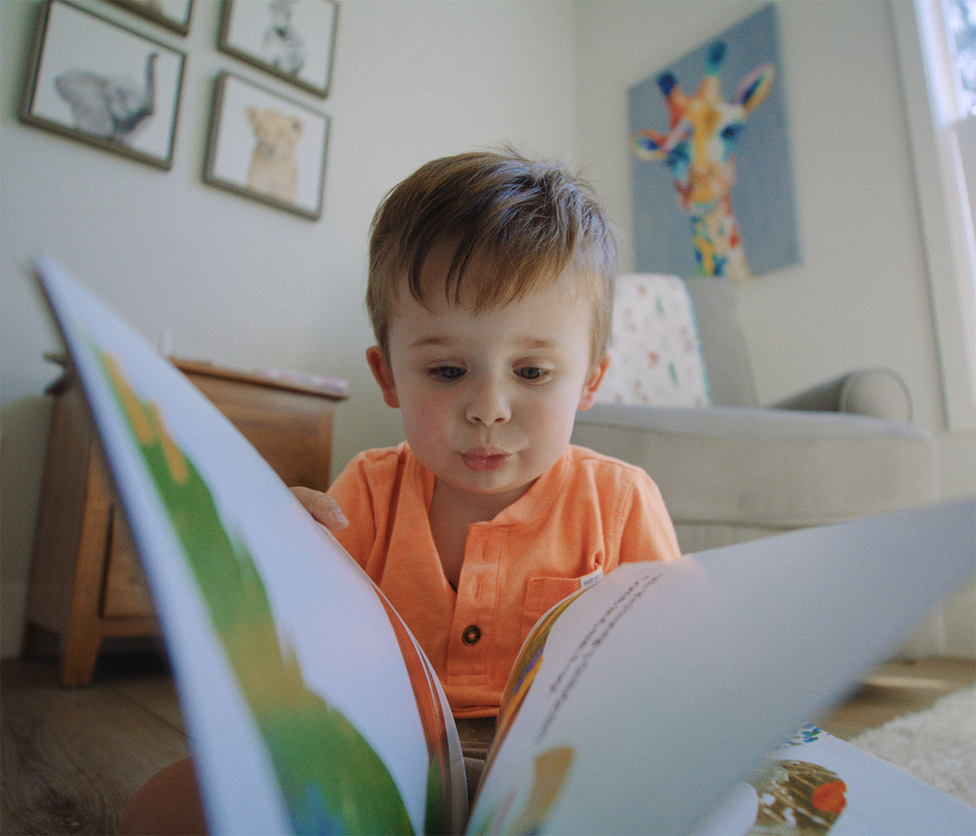 A toddler wearing a sunny orange shirt reads a book on the floor of his bedroom