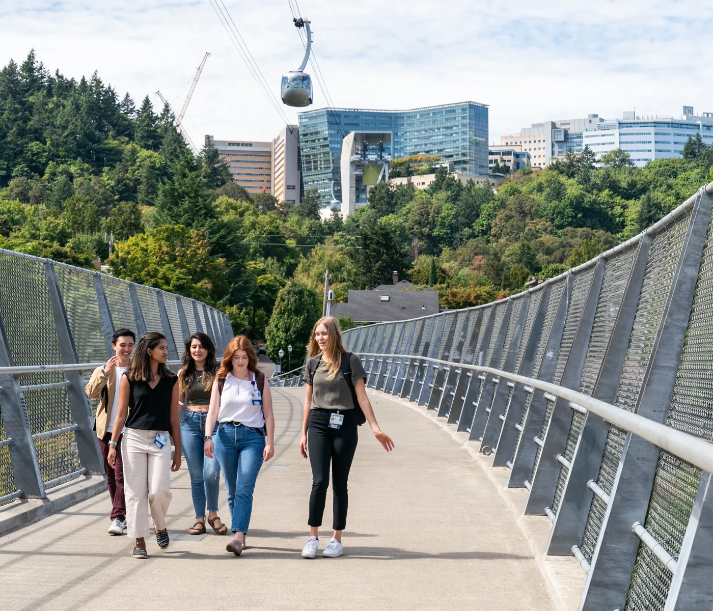 Five students walking across pedestrian bridge with OHSU Marquam HIll campus and aerial tram behind them.