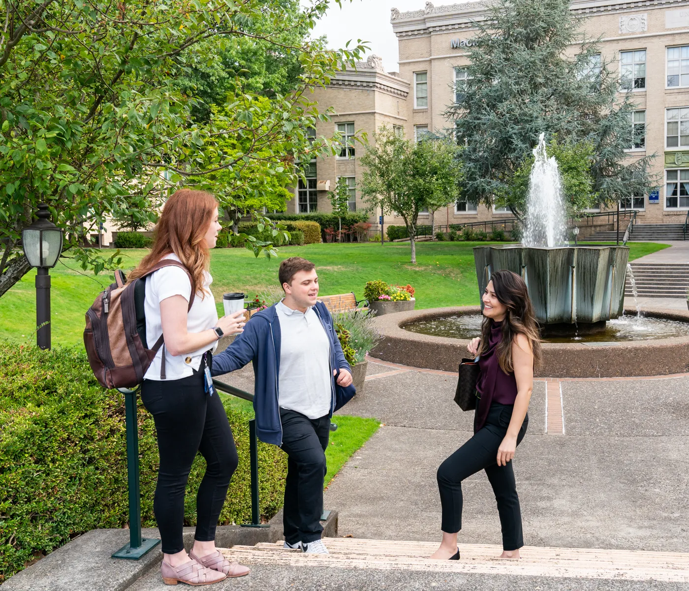 Three students stand on stairway talking outdoors with fountain and buildings behind them