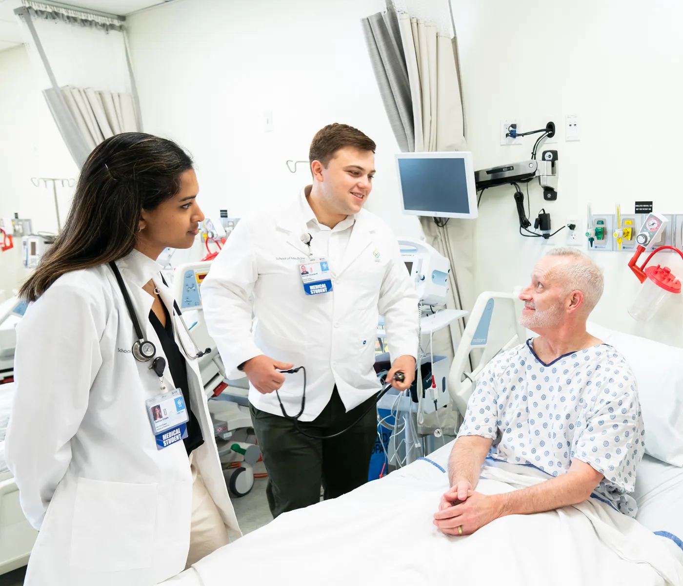 Two school of medicine students speak with male patient sitting up in bed.