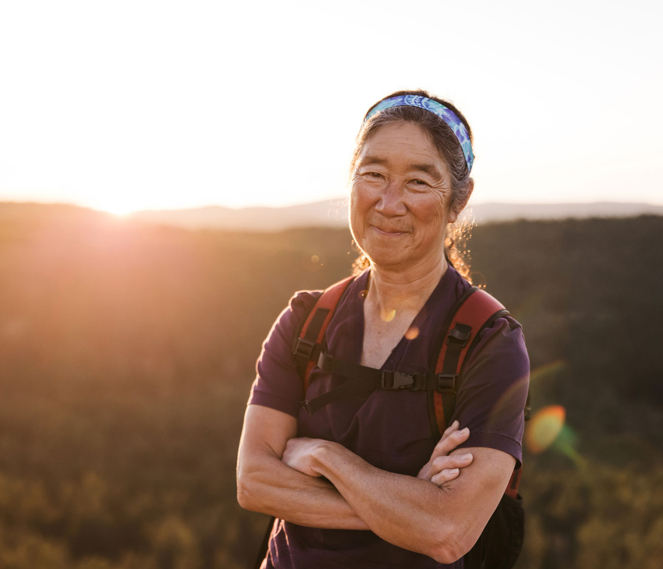 Mature woman wearing backpack stands outside with sun setting in distance