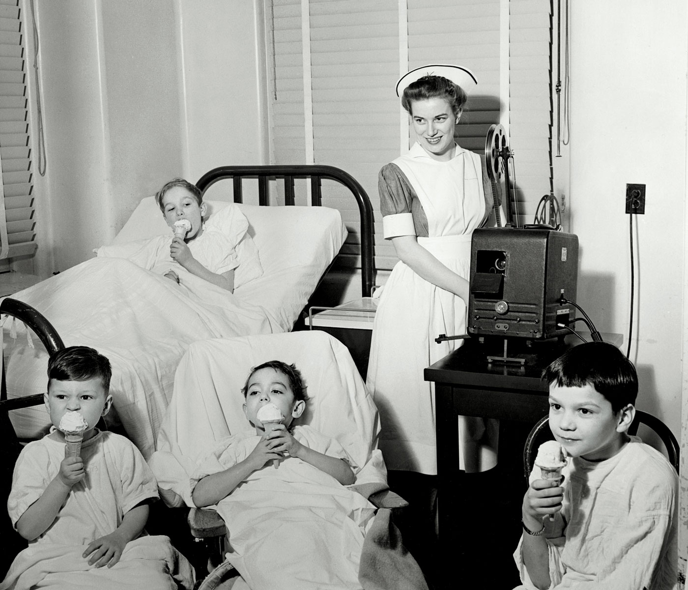 Kids in hospital gowns eat ice cream while nurse shows a movie on a projector, circa 1940