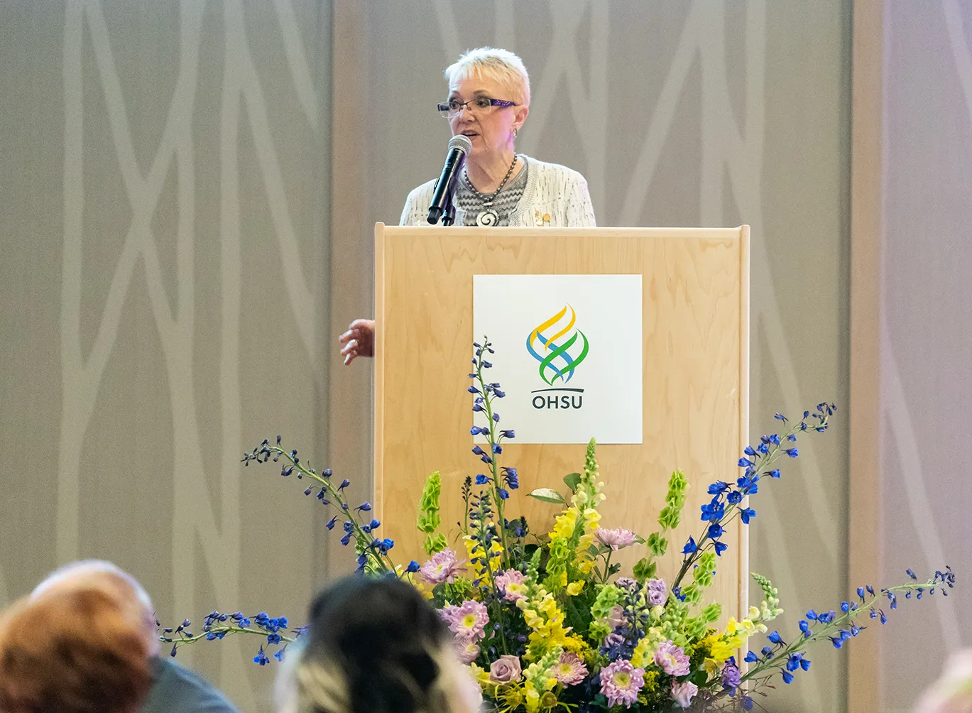 A women speaking at a podium with the OHSU logo on it and a large floral arrangement.