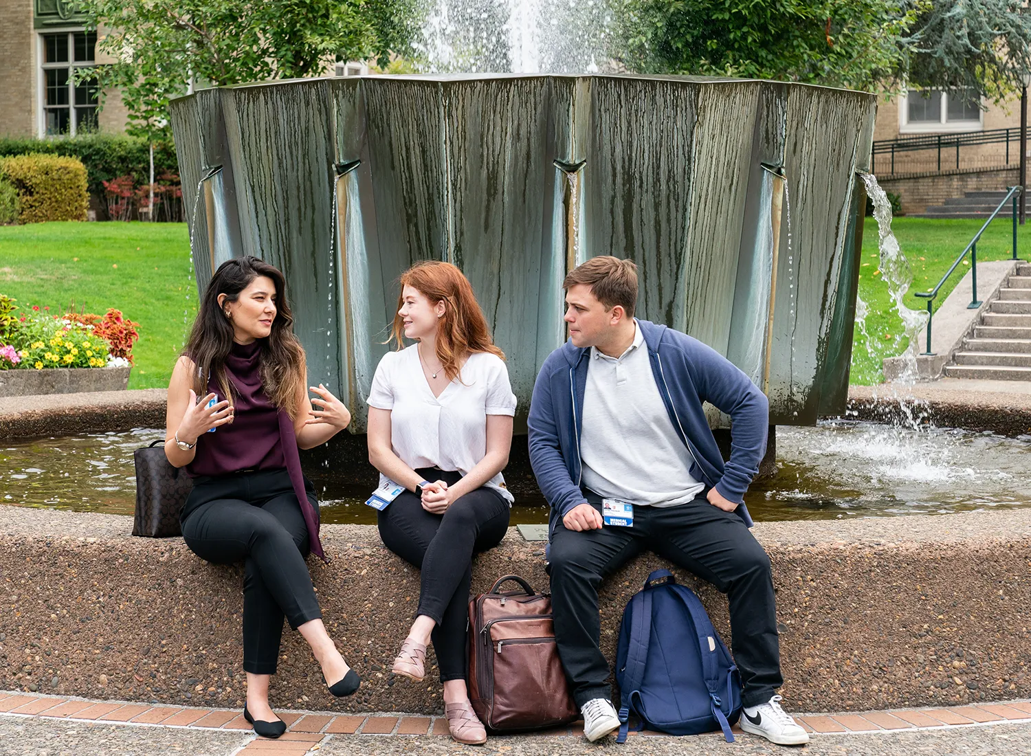 Two young women and a young man with book bags sitting side by side in front of a fountain.