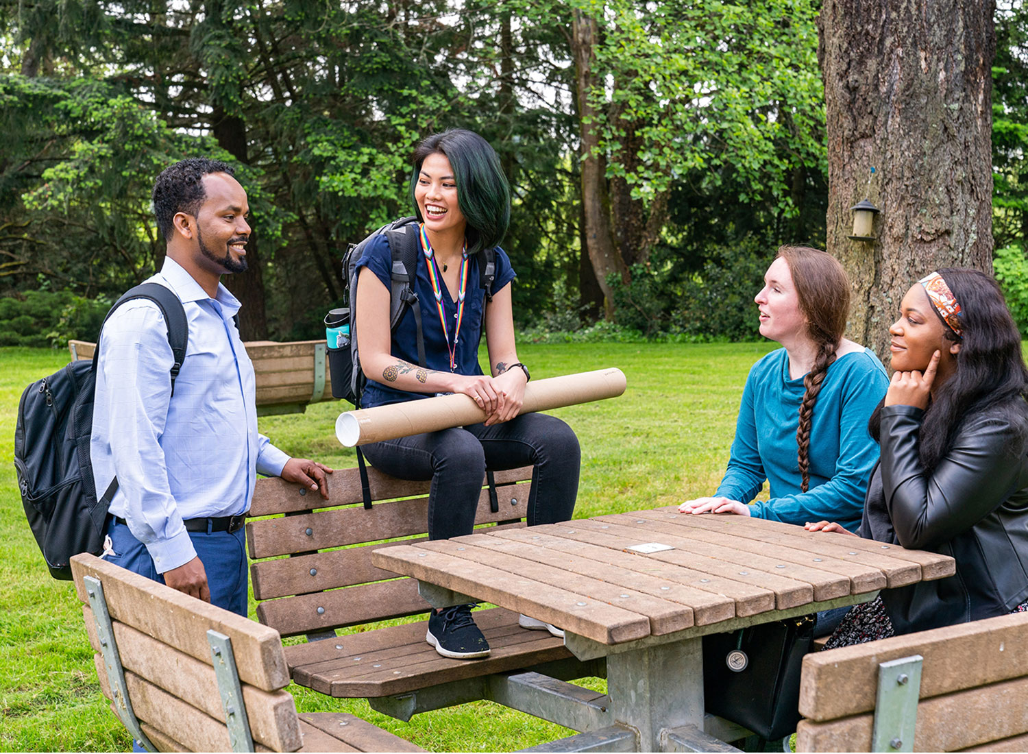 A young man wearing a backpack stands at a picnic table talking to three young women, one of whom is holding a cardboard poster tube.