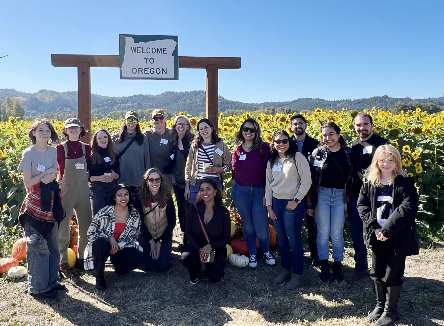 Group of PA alumni, smiling, standing outside on sunny day in front of sunflower field.