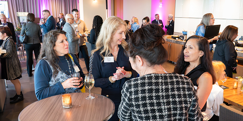 A group of women in business casual attire standing at a cocktail table talking and smiling.