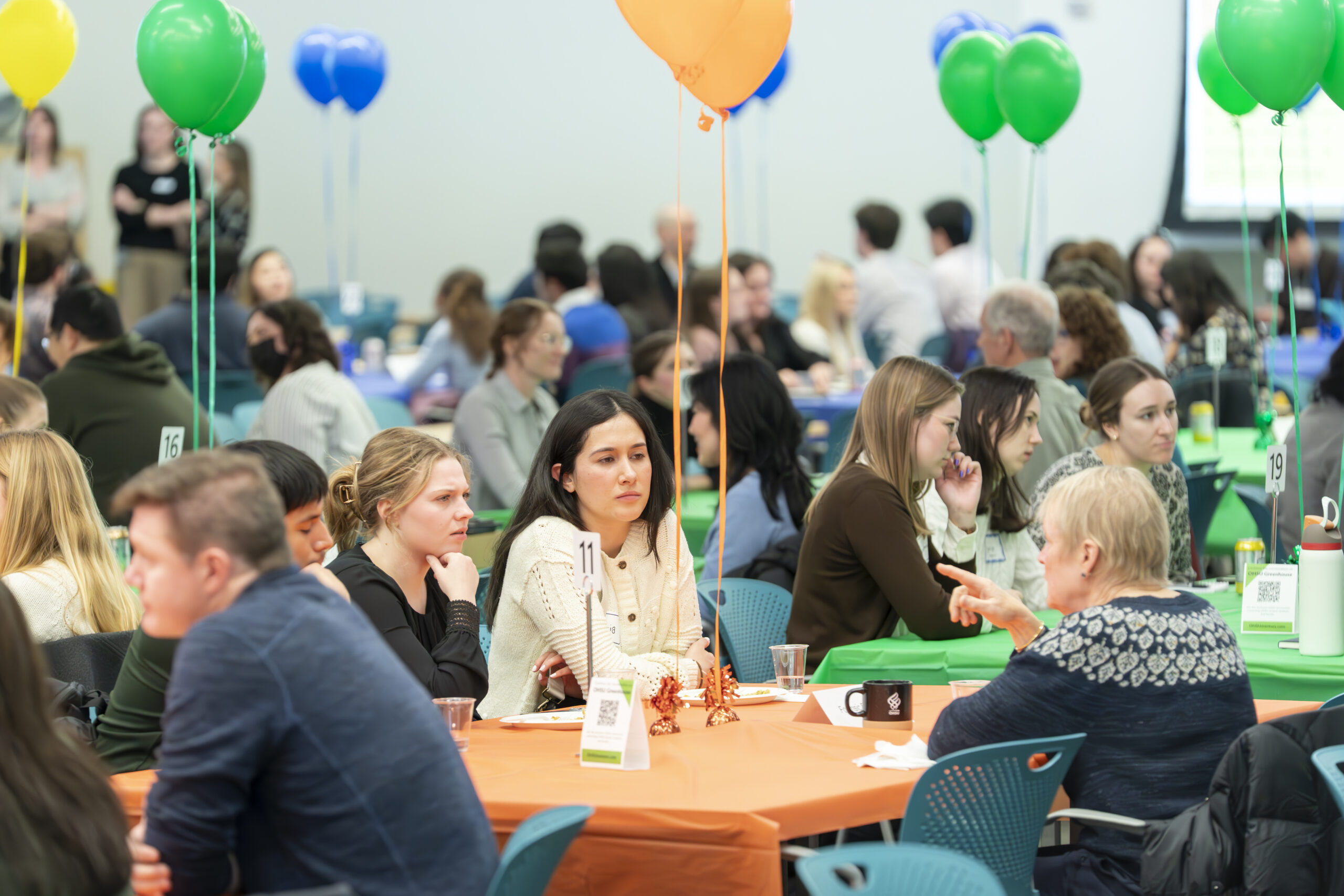 A group of people having lively conversations in a classroom with colorful tablecloths and balloons.
