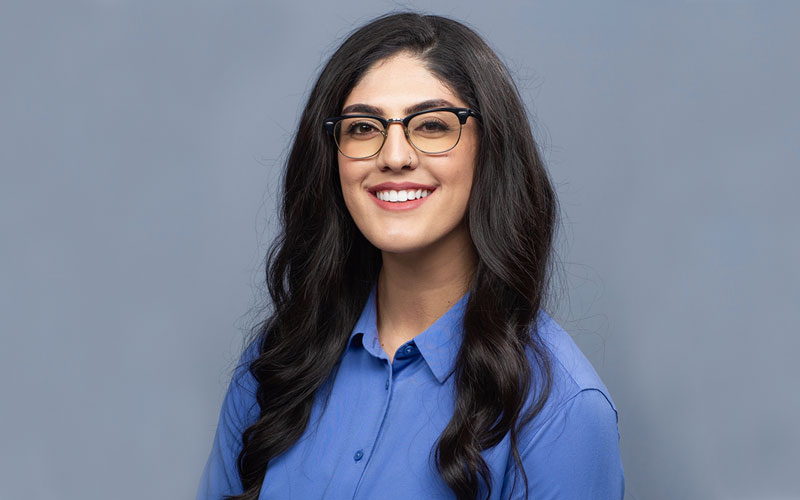 A headshot of a young woman with long, dark hair and glasses wearing a blue dress shirt