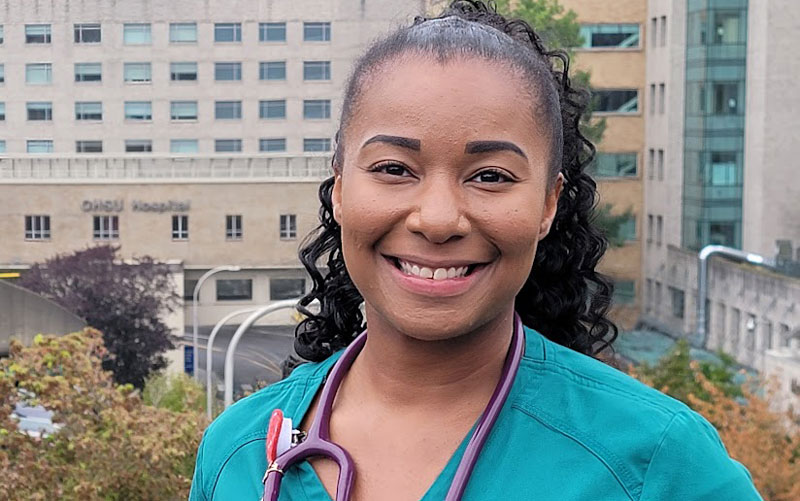 A young woman wearing turquoise scrubs and a stethoscope stands outside OHSU Hospital on Marquam Hill