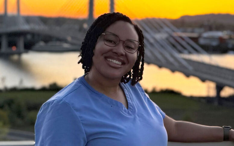 A young woman wearing blue scrubs and glasses stands with the Tilikum Bridge in the background at sunrise