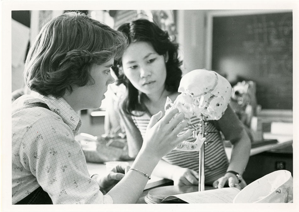A black and white photo of two young women in a classroom examining a model of a human skull.