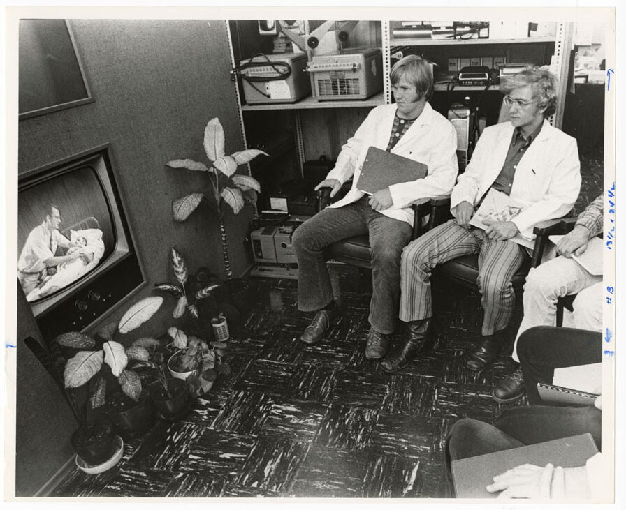 A small group of young men wearing white coats and holding clipboards sit in chairs in a semi-circle around an 1970s television showing a man leaning over a patient in a hospital bed.