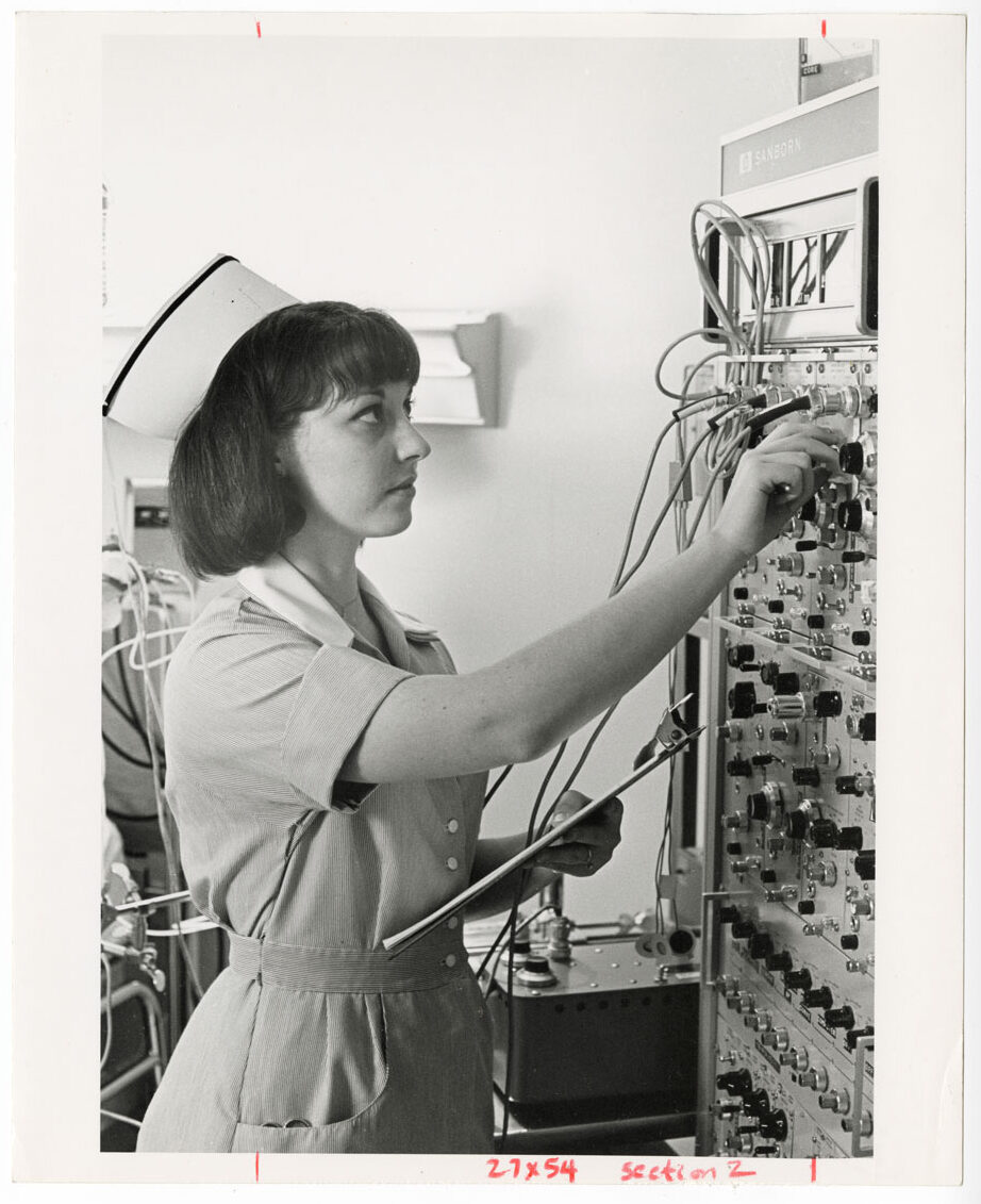 A black and white photo of a young woman in a dress and nurses cap holding a clipboard and touching the dials of a large machine.