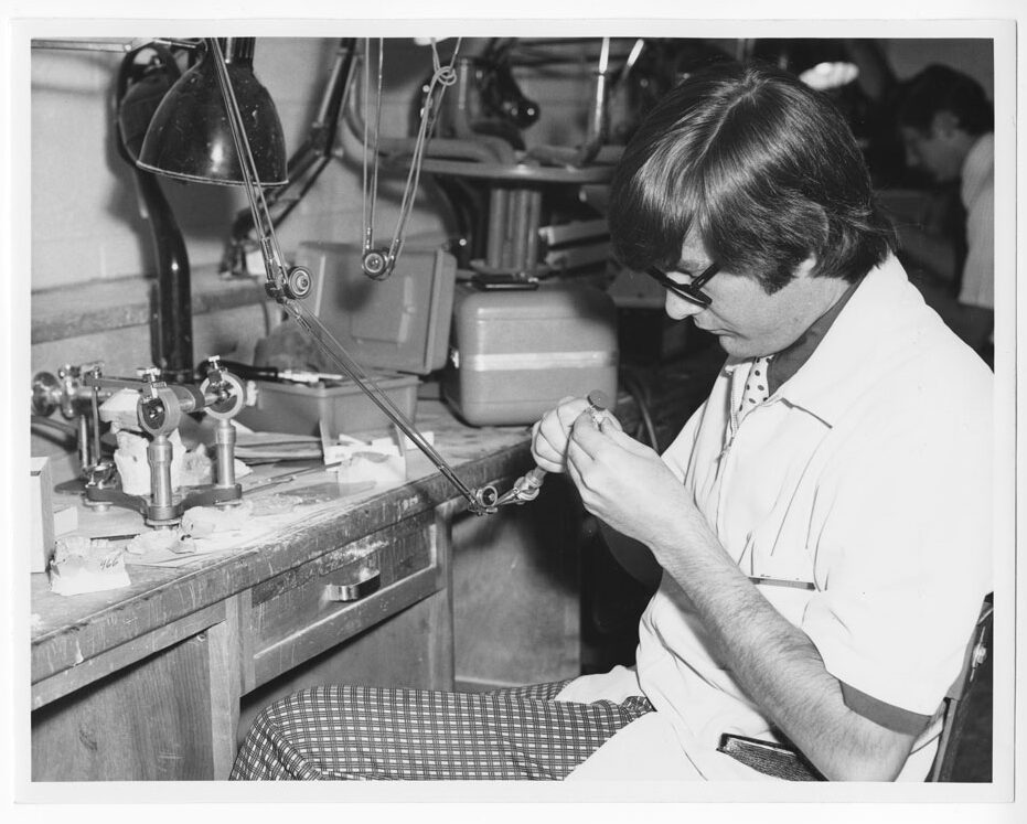 A black an white photo of a man wearing plaid pants and a polka dot tie looking down at a small tool in his hands.