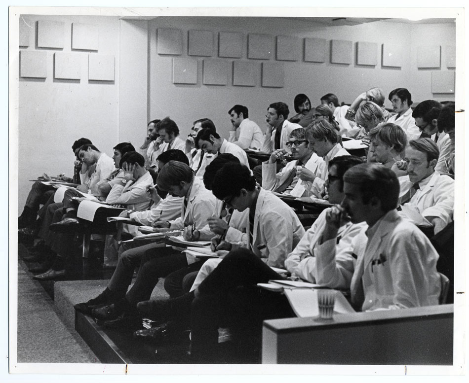 A black and white photo of a group of young men wearing white coats sitting in an auditorium-style classroom.