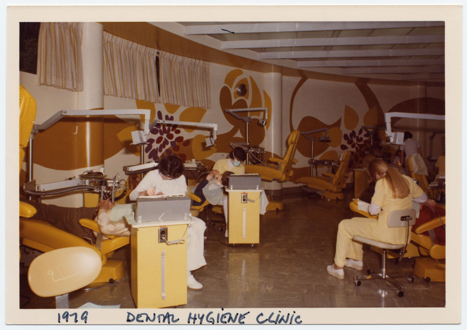 Three women lean over patients reclining in dental chairs in an clinic decorated with a 1970s-style mustard yellow and brown floral wall mural.