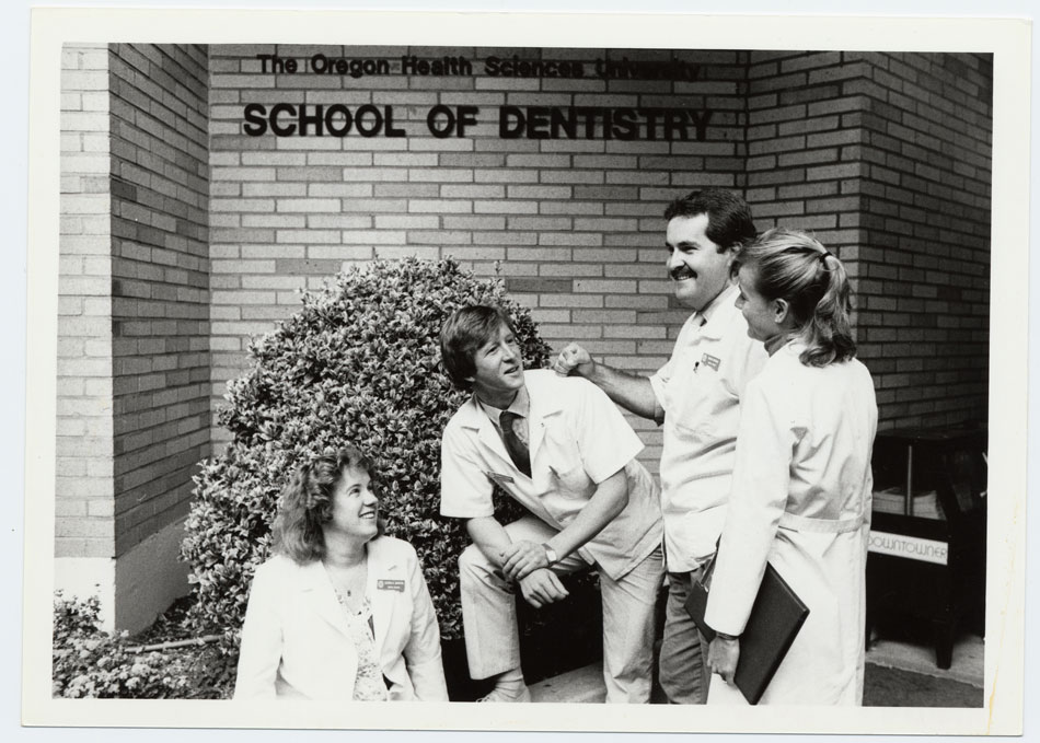 Two men and two women wearing white coats talk to each other and smile in front of a building with the words The Oregon Health Science University School of Dentistry on it.