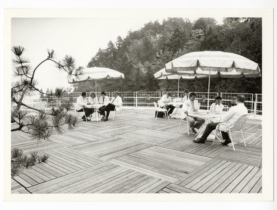 A group of men and women wearing white coats sitting at outdoor tables with umbrellas on a deck overlooking evergreen trees.