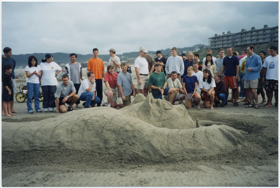 A group of young men and women standing on a beach near a large sand sculpture of a cat.