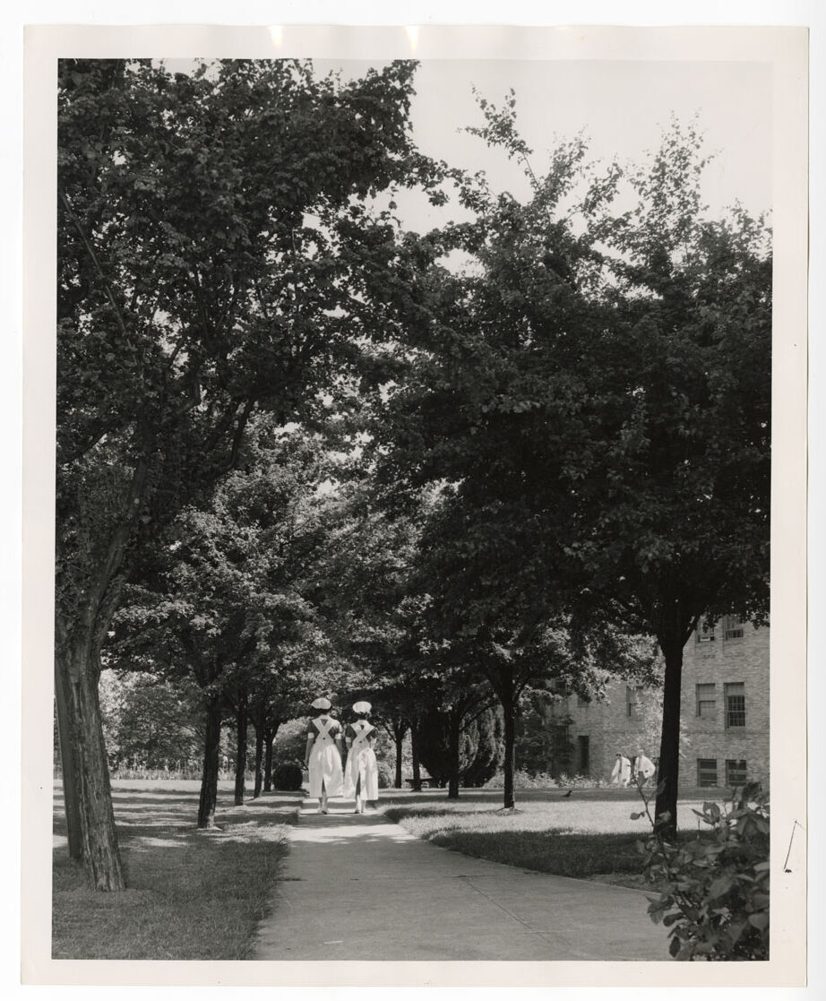 A black and white photo of two women wearing 1960s nursing uniforms walking on a tree-lined path.