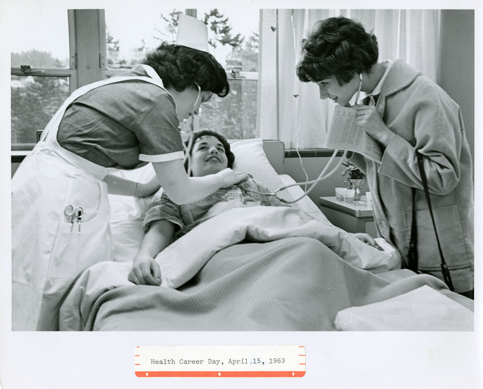 A black and white photo of two young women wearing stethoscopes leaning over a third young women lying in a hospital bed and smiling.