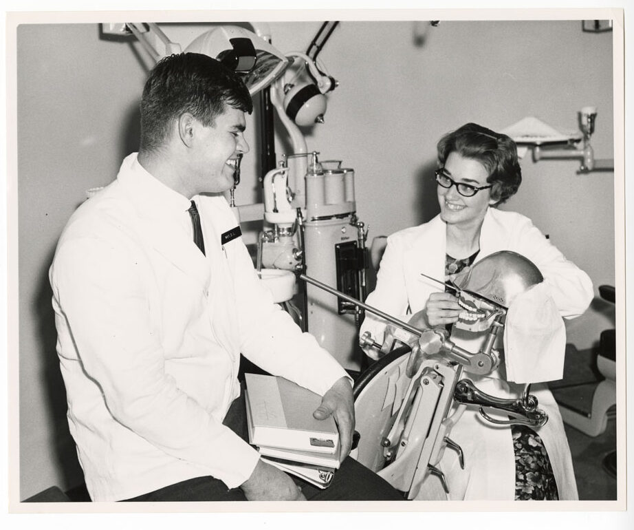 A black and white photo of a young man and a young woman with a dental training mannequin looking at each other and smiling.