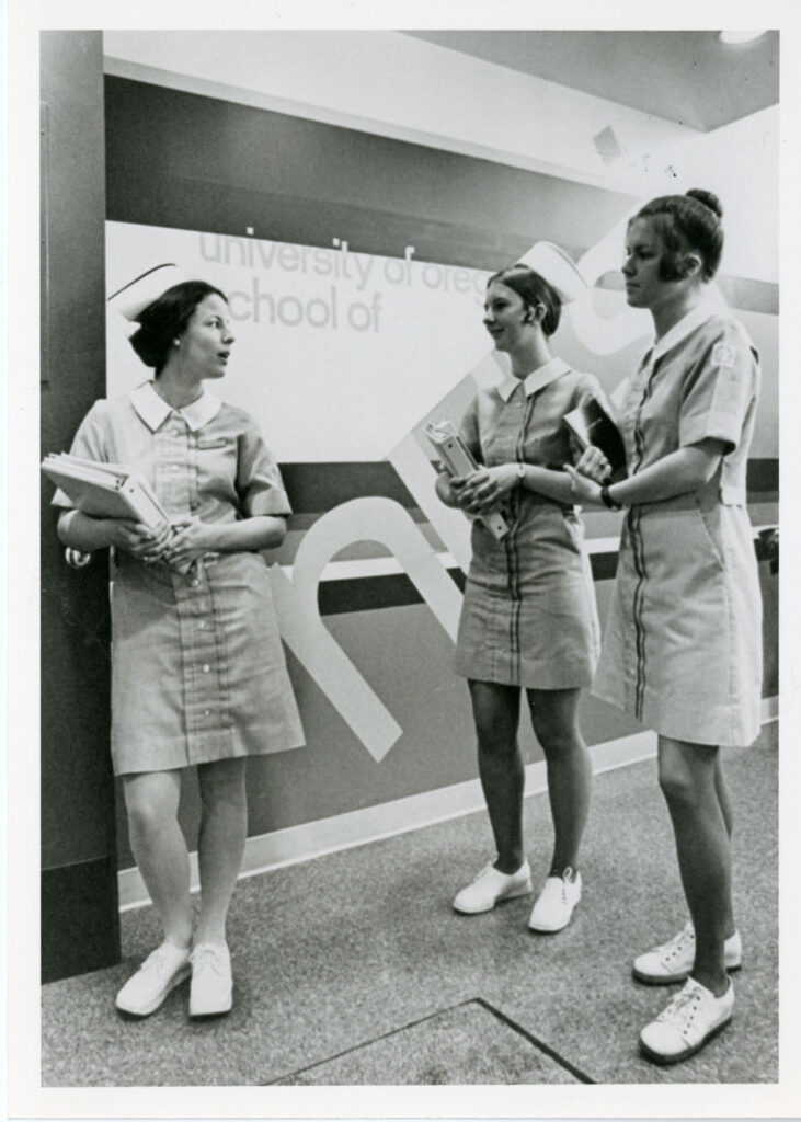 Three young women holding books and wearing 1960s nurses uniforms stand talking in a hallway.