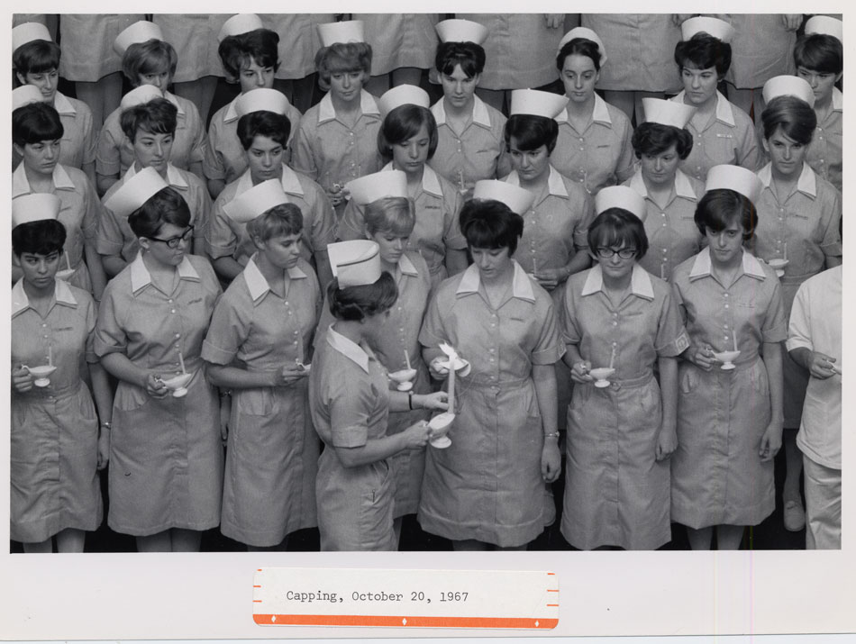 A black and white photo of a group of young women in 1960s nurses uniforms stand in rows holding candles.