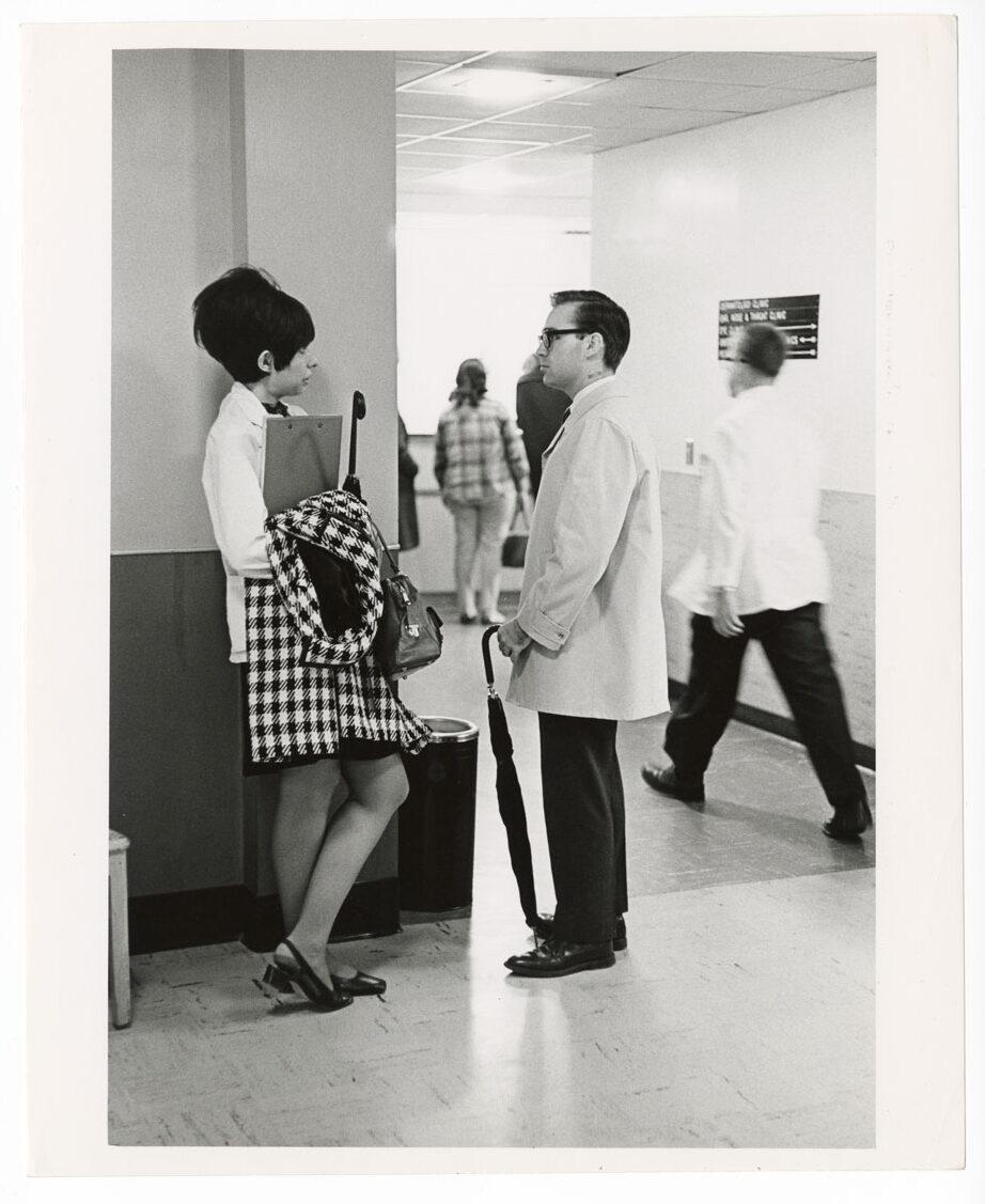 A young women wearing a white coat with a beehive hairstyle talks to a young man in a white coat and glasses holding an umbrella.