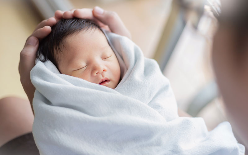 Infant with a full head of dark hair is held gently in a swaddling blanket