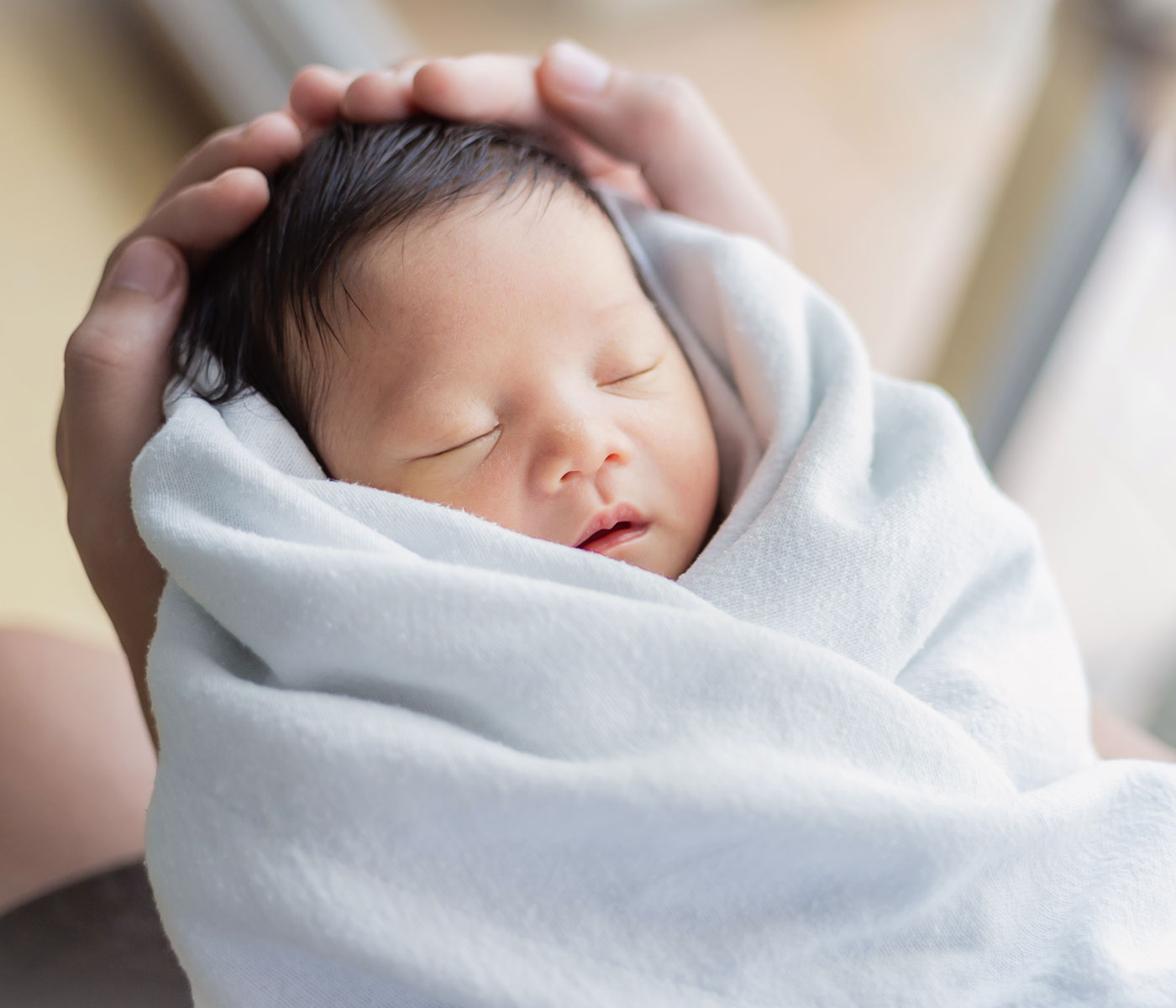 Infant with a full head of dark hair is held gently in a swaddling blanket