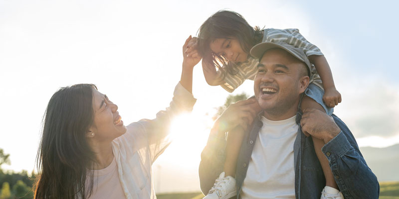 A family outdoors in the sunshine. A child rides on her father's shoulders.