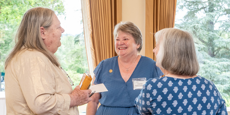 Three women stand in a sun-filled room smiling and talking.