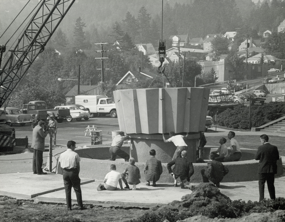 A black and white photo of men watching as a crane lowers a piece of the Alumni Fountain into place.