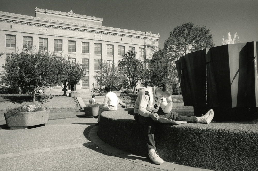 A young man in an argyle sweater sitting on a fountain bent over a book.